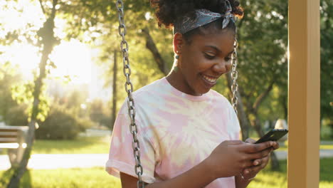 Joyous-African-American-Woman-Sitting-on-Swing-in-Park-and-Using-Phone